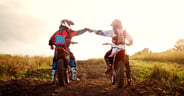 Rear view of dirt bikers bumping fists on field against sky